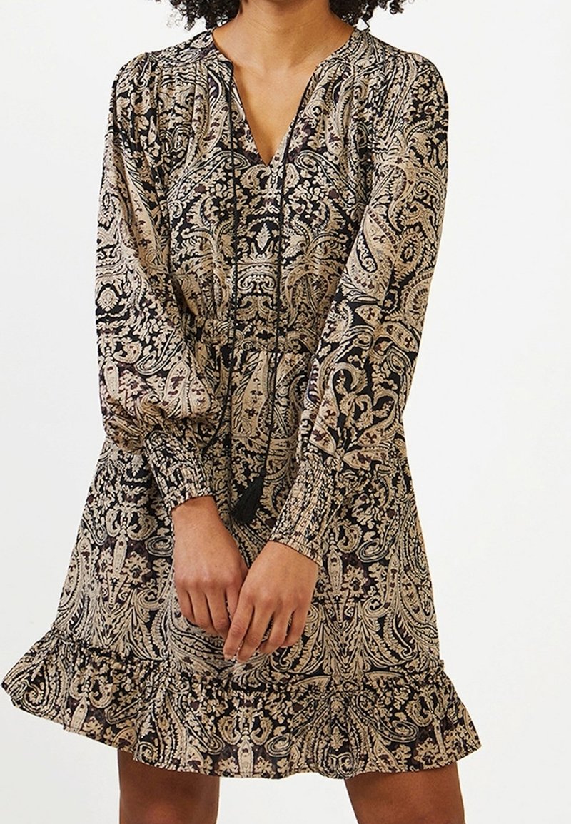 Femme portant une robe à manches longues, longueur genoux, avec un motif paisley beige et noir complexe et un ourlet à volants, mains jointes devant.