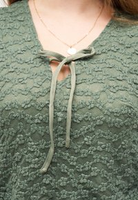 Κοντινή λήψη πράσινου δαντελένιου τοπ με λουλουδένια σχέδια, που διαθέτει δεμένο φιόγκο στη λαιμόκοψη και χρυσό στρογγυλό μενταγιόν κολιέ.