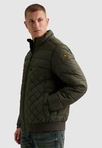 Homme portant une veste matelassée vert foncé avec un col montant et un patch sur la manche, debout avec une main dans la poche et regardant sur le côté.