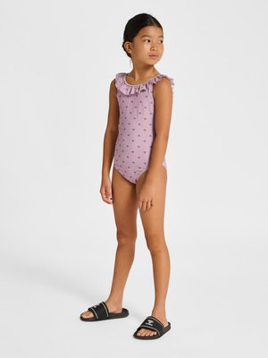 Junges Mädchen steht in lila Badeanzug mit gerüschtem Ausschnitt und kleinem Muster, trägt schwarze Slide-Sandalen und schaut seitlich vor weißem Hintergrund.