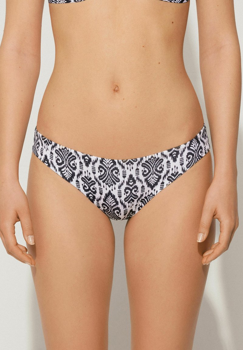 Bikiny bílé barvy s černým složitým vzorem. Materiál vypadá hladce, má nízký střih a minimální pokrytí látkou.