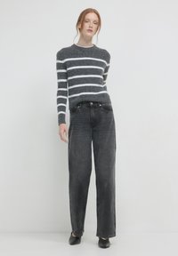 Une femme aux cheveux roux portant un pull rayé gris et blanc, un jean taille haute noir et des chaussures noires à bout pointu, se tenant devant un fond blanc.