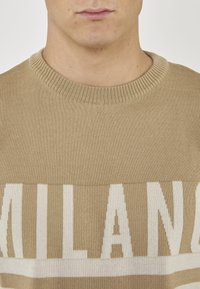 Maglione beige lavorato a maglia con scollatura rotonda, caratterizzato dalla parola "MILANO" in lettere bianche su tutto il petto. Texture liscia.