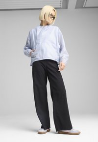 Veste bleu clair zippée en matériau lisse, associée à un pantalon large noir et des chaussures grises avec des accents clairs, sur un fond neutre.