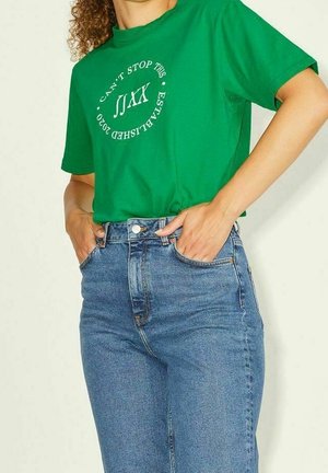 Kobieta ubrana w zieloną koszulkę z białym napisem i niebieskie jeansy z wysokim stanem, ręce schowane w przednich kieszeniach, na jednolitym tle.