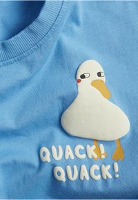 Sininen puuvillapaita, jossa on valkoinen ankka-graafika ja keltainen nokka sekä jalat. Alle on kirjoitettu "QUACK! QUACK!" valkoisilla kirjaimilla.