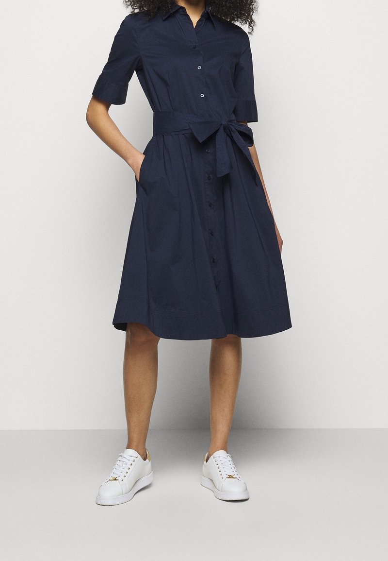 Modellen står i en marinblå klänning med knytmidja och fickor, iförd vita sneakers, mot en enkel grå bakgrund.