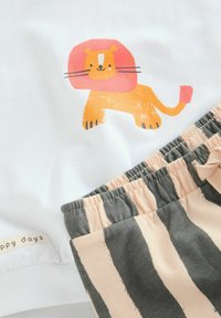 T-shirt blanche avec un graphisme de lion coloré et short beige à rayures grises. Comprend une taille élastique et une étiquette "jours heureux".