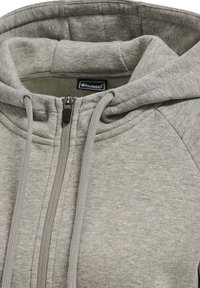 Sweat à capuche gris clair zippé avec cordons et col matelassé, montrant l'étiquette de la marque hummel à l'intérieur de la zone du cou.