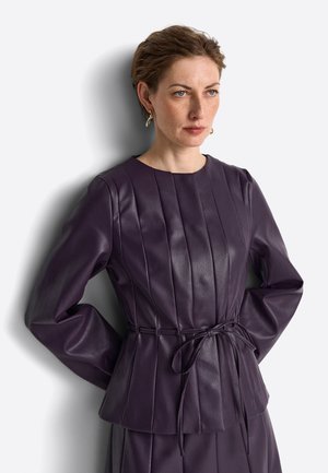 Frau mit kurzen Haaren, die eine eng anliegende dunkelviolette Lederjacke mit vertikalen Nähten und einem gebundenen Taillengürtel trägt, steht vor einem hellen Hintergrund.