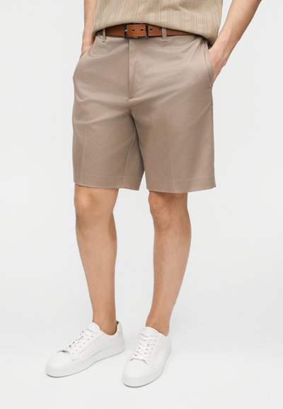Homme portant un short beige sur mesure avec une ceinture marron et des baskets blanches, les mains dans les poches, sur un fond uni.