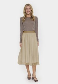 Jupe beige longueur cheville avec une silhouette fluide et une taille élastique, associée à un haut à manches longues de couleur marron clair et des ballerines à motifs.