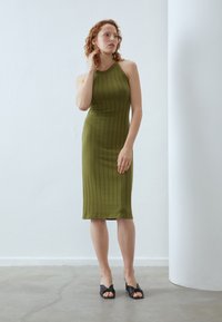 Olive green ribbed knit dress with a fitted silhouette, knee-length cut, and a high neckline; worn with black heeled sandals.