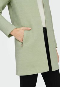 Abrigo verde claro con un patrón de espiguilla, que presenta un único bolsillo lateral y un escote estructurado, combinado con pantalones negros.