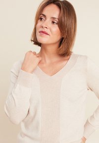Beige V-Ausschnitt-Pullover aus weichem Strickstoff, mit einem strukturierten Mittelteil und gerippten Bündchen für zusätzliche Details.