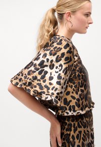 Satin topp med leopardmønster, brede ermer og volangkant. Har en miks av brune, gull og svarte toner. Modellen viser profil.