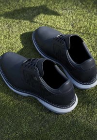 Chaussures de sport noires basses avec semelles blanches posées sur une surface d'herbe verte, vues de dessus et légèrement inclinées.