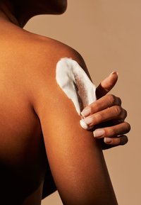 Ciało o gładkiej, opalonej skórze nakładające białą piankę na ramię, z naturalnym ułożeniem ręki i widocznymi jasnymi paznokciami.