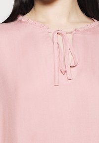 Blusa rosa realizzata in tessuto leggero con una sottile trama a coste, un colletto arricciato e un dettaglio a chiave legato sul davanti.
