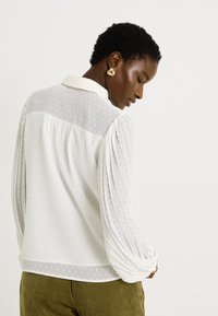 Blusa blanca con lunares texturizados, mangas largas plisadas y cuello con solapa. La tela parece ligera y semitranslúcida, combinada con pantalones verdes.