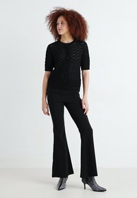 Cardigan noir en maille à manches courtes avec un motif en diamant texturé, associé à un pantalon noir évasé près du corps et des bottes à talons brillantes.