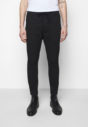 Persona vistiendo pantalones ajustados negros con cintura de cordón y botas negras hasta el tobillo, de pie frente a un fondo blanco.