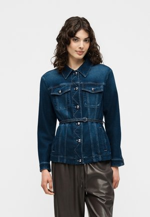 Frau mit lockigem Haar, die eine dunkelblaue Jeansjacke mit Knöpfen und einem Taillengürtel trägt, kombiniert mit dunkelbraunen Lederhosen.