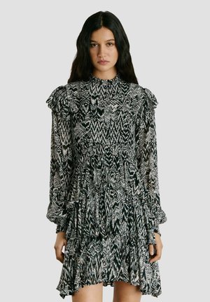 Vrouw met lang donker haar, gekleed in een zwarte en witte jurk met hoge hals en lange mouwen, staand tegen een effen achtergrond.