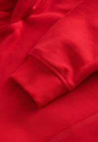 Der Stoff des roten Kapuzenpullovers zeigt eine glatte Textur und hat einen gerippten Ärmelbund. Der Bund ist etwas dunkler und elastischer als der Rest des pullovers.
