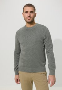 Homme aux cheveux courts portant un pull gris à manches longues et un pantalon beige, debout devant un fond clair uni.