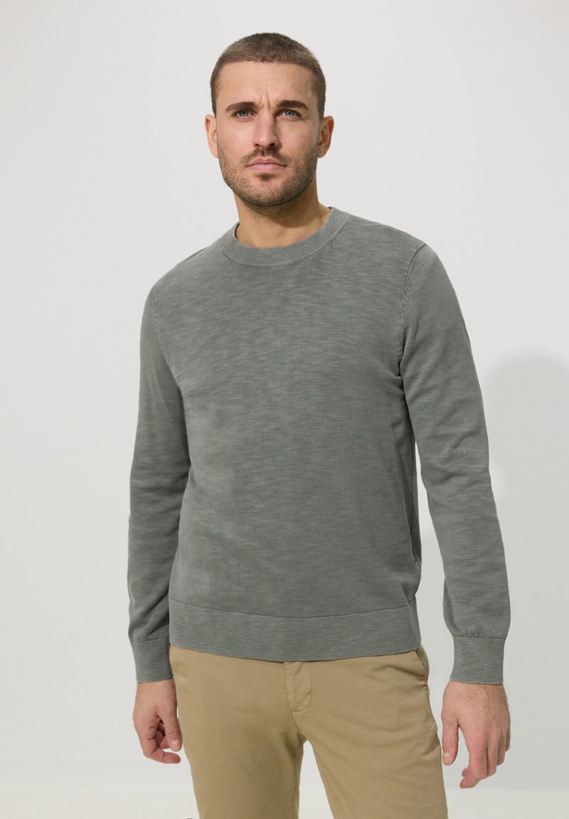 Homme aux cheveux courts portant un pull gris à manches longues et un pantalon beige, debout devant un fond clair uni.