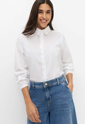 Lächelnde Frau mit langen dunklen Haaren, die eine weiße Bluse mit Rüschen und blaue High-Waist-Jeans trägt, die Hände in den Taschen.