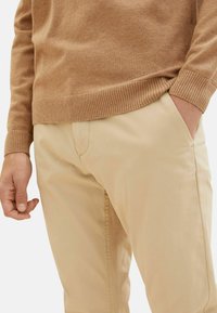 Ljusbrun stickad tröja med ribbad mudd, kombinerad med beige byxor som har en sidficka och en jämn textur.
