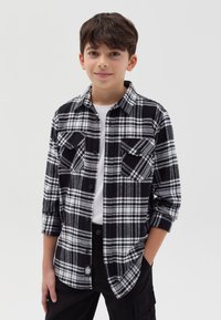 Camisa de franela a cuadros en blanco y negro con mangas largas, dos bolsillos frontales y cierre de botón; llevada sobre una camiseta blanca y pantalones negros.