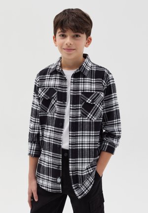 Camisa de franela a cuadros en blanco y negro con mangas largas, dos bolsillos frontales y cierre de botón; llevada sobre una camiseta blanca y pantalones negros.