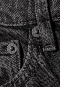 Close-up al unui material din denim negru, prezentând un buton metalic cu cuvântul "WEEKDAY," o cusătură vizibilă și un finisaj texturat, întunecat.