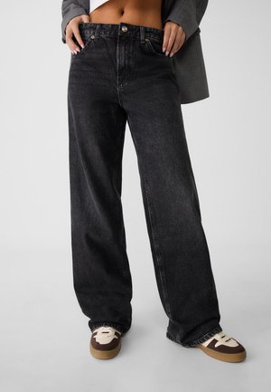 Jean large noir taille haute avec texture délavée, doté de poches avant et d'une fermeture à bouton métallique, associé à des baskets marron.
