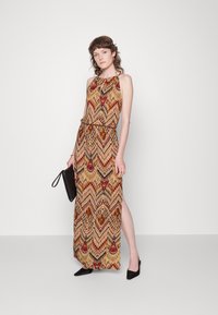 Maxikleid mit Halterneck, mehrfarbigem Zickzackmuster in erdigen Tönen, tailliert und mit seitlichem Schlitz. Ergänzt durch eine schwarze Clutch.