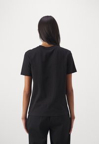 Kvinna med långt mörkt hår iförd en enkel svart t-shirt med kort ärm och svarta byxor, stående vänd bort mot en vit bakgrund.