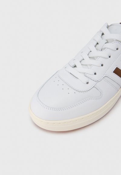 Λευκό δερμάτινο αθλητικό παπούτσι με στρογγυλεμένη μύτη, διάτρητες λεπτομέρειες, επίπεδα κορδόνια και καφέ διακοσμητικό στη πλευρά. Λαστιχένια σόλα με ελαφρώς κρεμ απόχρωση.