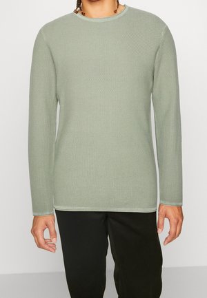 Chemise longue à manches longues vert clair en tissu texturé avec un col rond. Les poignets et l'ourlet taillés ajoutent du détail. Portée avec un pantalon noir.