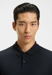 Svart pikétröja i texturerad väv; har en klassisk krage, två knappar på placket och korta ärmar, vilket ger en figurnära silhuett.