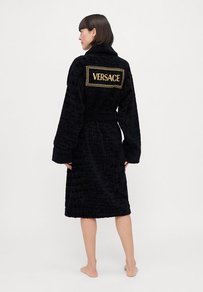 Robe en velours noir avec un motif texturé, des manches larges et une taille ceinturée. Le dos présente un logo doré "VERSACE" bien en vue.