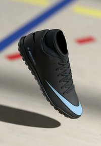 Scarpa da calcio Nike Mercurial nera con swoosh e lacci blu, mostrata in volo sopra un campo al coperto con linee colorate.