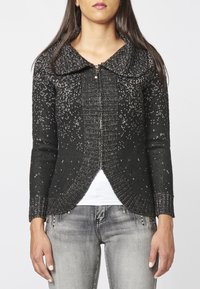 Black knitted cardigan with a rounded collar, features silver specks and a front zipper. Textured finish and fitted cuffs. Worn over a white top.