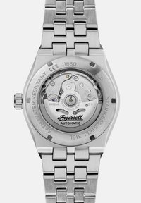 Montre automatique en acier inoxydable argenté avec un dos hexagonal, montrant des détails de mouvement complexes et un marquage gravé. Fini brossé doux.