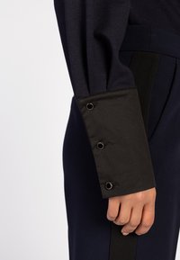 Marineblauses Langarmshirt mit einem schwarzen Bündchen, das drei dekorative Metall-Ösen aufweist. Der Stoff hat eine glatte Textur mit einem dezenten Glanz.