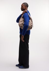 Beige stoffen schoudertas met een zachte textuur, trekkoordsluiting en een ruim ontwerp, gedragen op de schouder door een persoon in een blauwe trui.