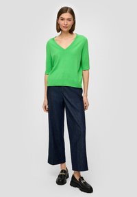 Grünes V-Ausschnitt-Stricktop mit kurzen Ärmeln, kombiniert mit lockeren, dunklen Jeans. Schwarze Slipper mit goldenen Akzenten runden den Look ab.