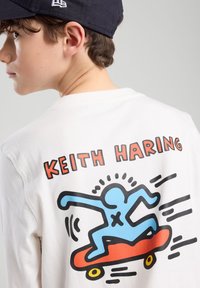 T-shirt à manches longues de couleur blanche avec un graphique coloré de skateboard de Keith Haring. Tissu en coton doux avec un col rond.